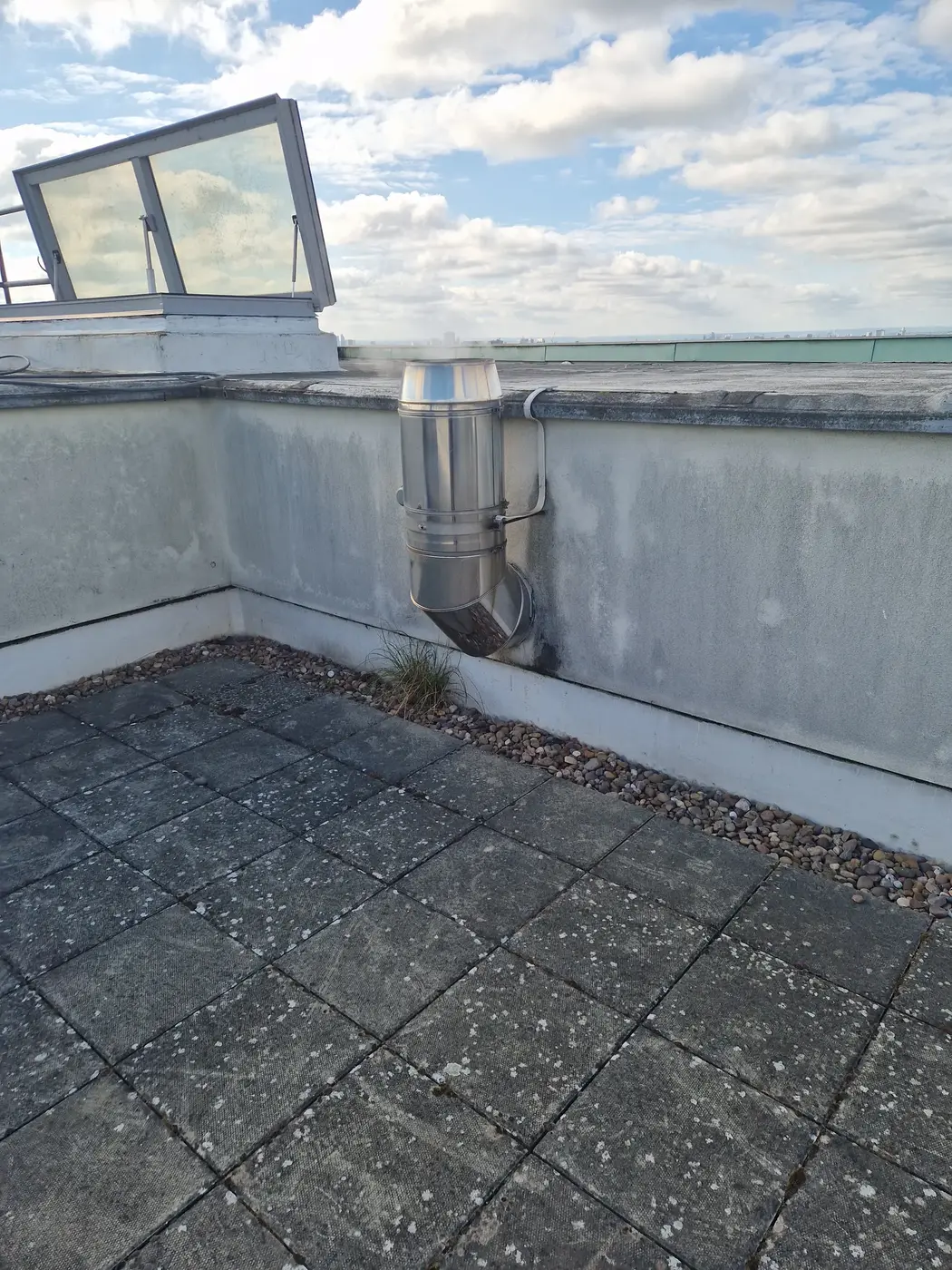A flue terminal on a flat roof terrace, next to the parapet wall and a roof hatch